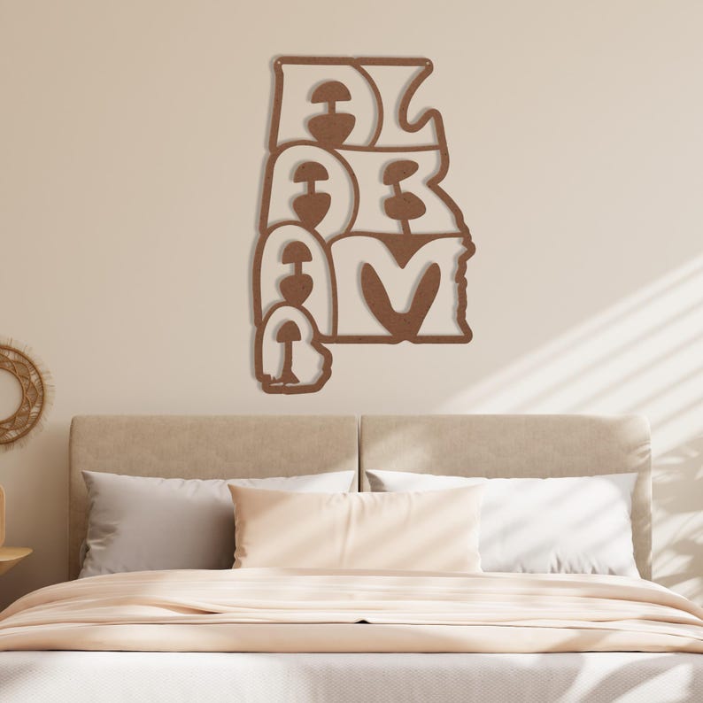 Puede incluir: Adorno de pared de madera con la palabra "ALABAMA" escrita en una fuente apilada y estilizada. Las letras son de color marr&oacute;n c&aacute;lido, con un contorno marr&oacute;n m&aacute;s claro. La obra de arte est&aacute; montada en una pared de color neutro sobre una cama.