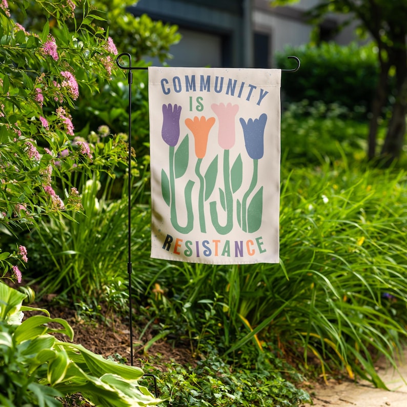 May include: A white garden flag with the message "Community is Resistance" in colourful letters. The flag features a stylised design of four tulips in purple, orange, pink, and blue.