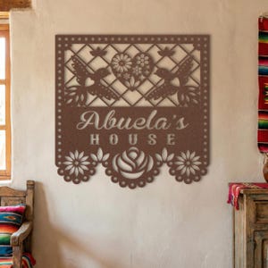 Op de afbeelding: Een bruin metalen wandkunstwerk met een decoratief uitgesneden ontwerp. De kunst bevat de woorden "Abuela's House" en bevat afbeeldingen van vogels, bloemen en een hart. Het stuk is ontworpen om aan een muur te worden gehangen.