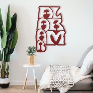 Puede incluir: Una pieza de arte mural de metal rojo que deletrea "ALABAMA" en una fuente estilizada. La obra de arte est&aacute; montada en una pared blanca, con un sof&aacute; y plantas en el fondo. La pieza mide aproximadamente 61 cm de ancho y 91 cm de alto.