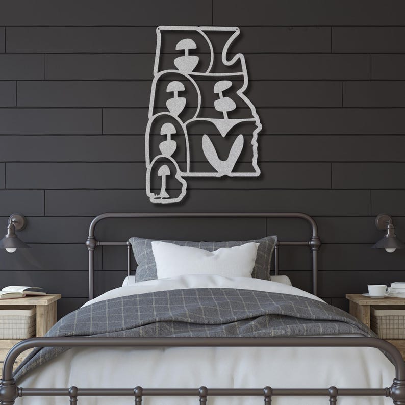 Puede incluir: Una pieza de arte mural de metal plateado que deletrea la palabra "ALABAMA" en una fuente estilizada. La obra de arte est&aacute; montada en una pared de madera gris oscuro sobre una cama con un edred&oacute;n gris y blanco.