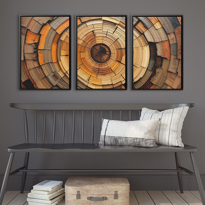 May include: Three framed abstract art pieces depicting cross-sections of wood, showcasing concentric circles in shades of brown, orange, and tan. The artwork is displayed above a gray bench with pillows and a suitcase.