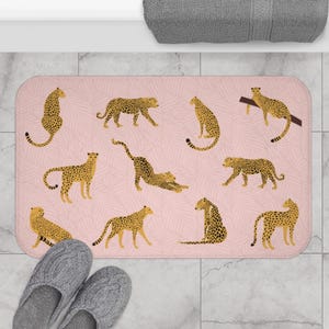 May include: A pink bath mat with a repeating pattern of cheetah print. The cheetah print is in shades of brown and yellow. The mat is rectangular and has a textured surface.