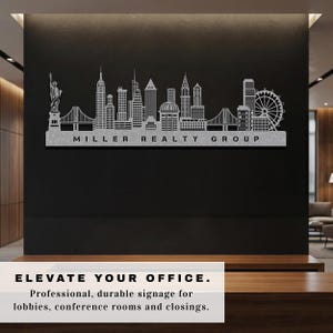 May include: A silver metal skyline silhouette featuring the Statue of Liberty, bridges, and skyscrapers. The text "MILLER REALTY GROUP" is below the skyline. The image is mounted on a black wall. The text "ELEVATE YOUR OFFICE" is at the bottom.