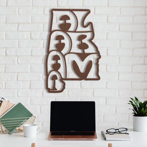 Puede incluir: Una pieza de arte mural de metal marr&oacute;n que deletrea la palabra "DREAM" en una fuente estilizada. La obra de arte est&aacute; montada en una pared de ladrillo blanco. Una computadora port&aacute;til, una taza de caf&eacute; y otros suministros de oficina est&aacute;n en un escritorio blanco debajo.