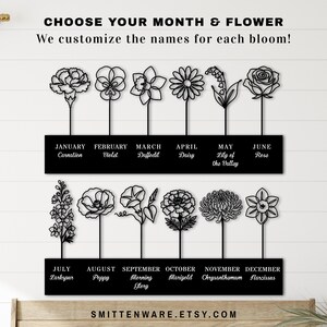 K&ouml;nnte beinhalten: Schwarze Metall-Wandkunst mit stilisierten Blumendesigns, die jeden Monat des Jahres darstellen. Jede Blume ist mit dem entsprechenden Monat und Namen beschriftet. Der Text "CHOOSE YOUR MONTH & FLOWER" steht oben.