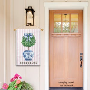 May include: A personalized garden flag with a pink and white striped background. A blue and white porcelain planter with a green topiary tree and a blue bow is in the center. The text "ROBERTSON" is printed below the planter.