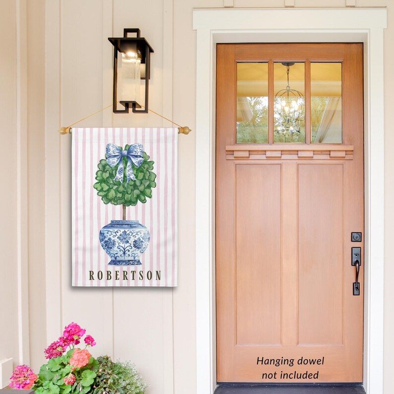 May include: A personalized garden flag with a pink and white striped background. A blue and white porcelain planter with a green topiary tree and a blue bow is in the center. The text "ROBERTSON" is printed below the planter.