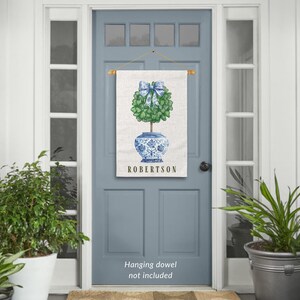 May include: A personalized door hanger with a blue and white porcelain pot with a green topiary tree and a blue bow. The text "ROBERTSON" is printed on the door hanger.