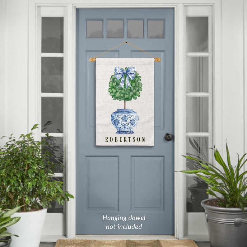 May include: A personalized door hanger with a blue and white porcelain pot with a green topiary tree and a blue bow. The text "ROBERTSON" is printed on the door hanger.