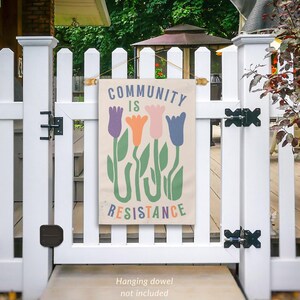 May include: A beige canvas banner with the text "COMMUNITY IS RESISTANCE" in colourful letters. The banner features a stylised design of four colourful tulips with green stems and leaves.