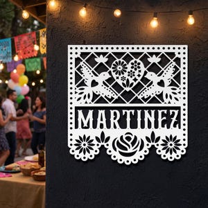 Op de afbeelding: Witte lasergesneden wandkunst met de naam "MARTINEZ". Het ontwerp toont vogels, bloemen en een bloemstuk in de vorm van een hart. De onderste rand heeft een geschulpte bloemenrand. De kunst is op een donkere muur gemonteerd.