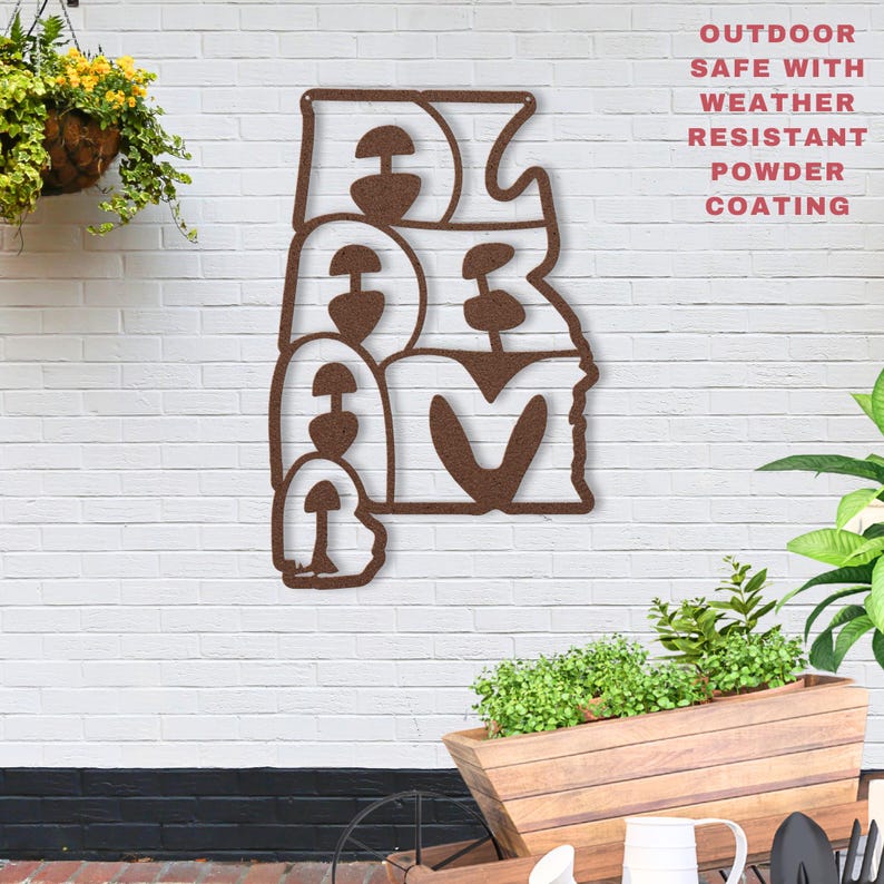 Puede incluir: Una pieza de arte mural de metal marr&oacute;n que deletrea la palabra "DREAM" en una fuente estilizada. La obra de arte est&aacute; montada en una pared de ladrillo blanco, con una cesta colgante de flores amarillas y una jardinera de madera con vegetaci&oacute;n en primer plano.