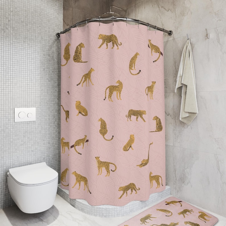 May include: A pink shower curtain with a pattern of gold leopards. The shower curtain is in a bathroom with a white toilet and a white towel hanging on a hook.