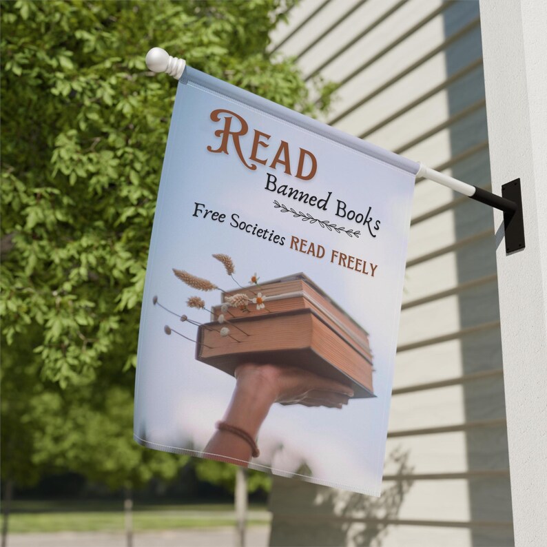 Może przedstawiać: Biała flaga z tekstem "READ Banned Books Free Societies READ FREELY" i ręką trzymającą stos książek.