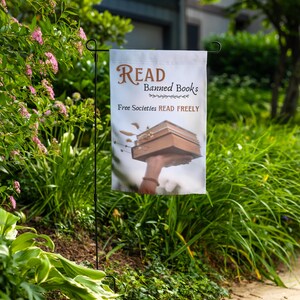 Może przedstawiać: Biała flaga ogrodowa z tekstem "Read Banned Books" i "Free Societies Read Freely". Flaga przedstawia rękę trzymającą stos książek.