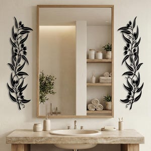 May include: Black metal wall art featuring olive branch designs. The art pieces are installed on either side of a large mirror in a bathroom setting. The olive branches have leaves and small, round fruit.