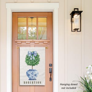 May include: A personalized door flag with a blue and white porcelain planter with a green topiary tree and a blue bow. The flag is white with a linen texture and the text "ROBERTSSON" is printed in black at the bottom.