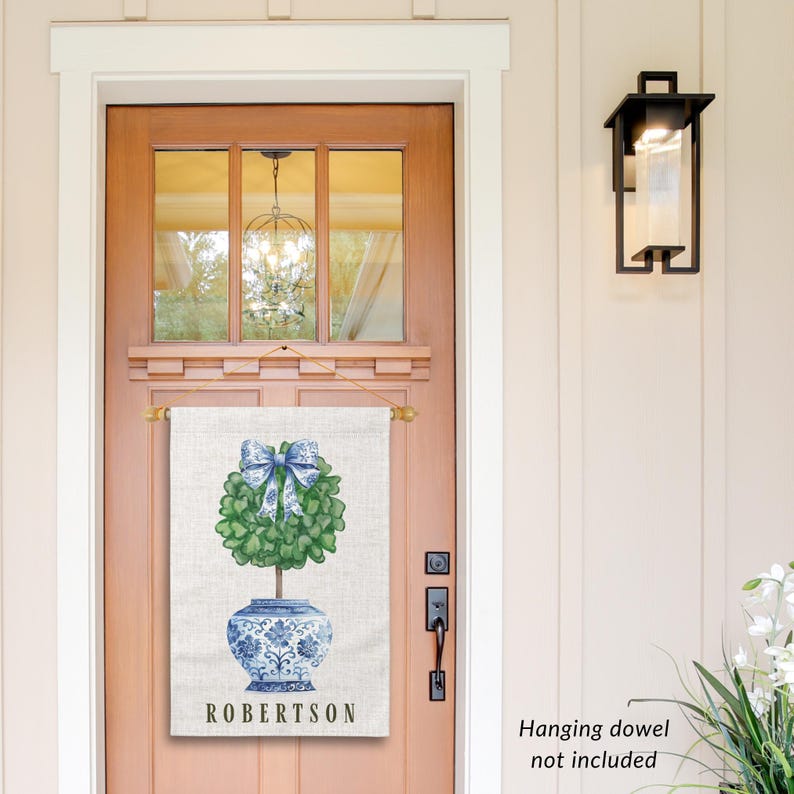 May include: A personalized door flag with a blue and white porcelain planter with a green topiary tree and a blue bow. The flag is white with a linen texture and the text "ROBERTSSON" is printed in black at the bottom.