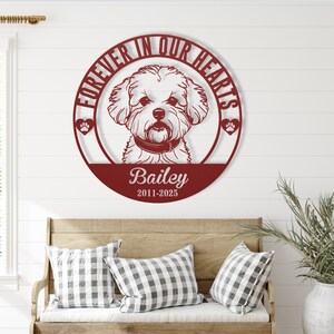 Puede incluir: Una pieza de arte mural de metal burdeos con un retrato de perro dentro de un marco circular. El texto "FOREVER IN OUR HEARTS" se arquea sobre el perro, con el nombre "Bailey" y las fechas "2011-2025" debajo. Huellas de patas flanquean la cabeza del perro.