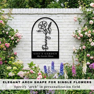 K&ouml;nnte beinhalten: Schwarzes Metall-Gartenschild mit einem bogenf&ouml;rmigen Rahmen, der ein Blumendesign und den Namen "Danny" zeigt. Das Schild enth&auml;lt auch den Text "GIGI'S GARDEN My Favorite Bloom". Das Schild ist an einer wei&szlig;en Backsteinmauer angebracht, umgeben von Blumen.