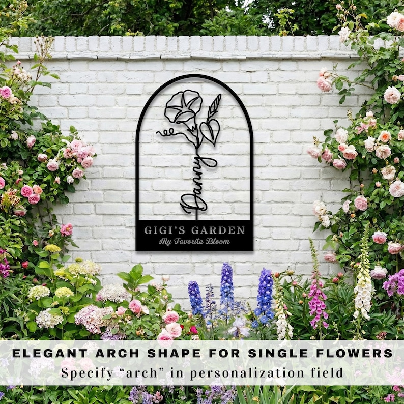 K&ouml;nnte beinhalten: Schwarzes Metall-Gartenschild mit einem bogenf&ouml;rmigen Rahmen, der ein Blumendesign und den Namen "Danny" zeigt. Das Schild enth&auml;lt auch den Text "GIGI'S GARDEN My Favorite Bloom". Das Schild ist an einer wei&szlig;en Backsteinmauer angebracht, umgeben von Blumen.