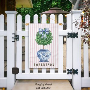 May include: A pink and white striped garden flag with a blue and white porcelain vase with a green topiary tree and a blue bow. The flag has the name "ROBERTSON" printed on it.