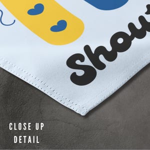 K&ouml;nnte beinhalten: Ein wei&szlig;es Stoffbanner mit blauen und gelben Formen und dem Text "Baby Shower" in Schwarz.