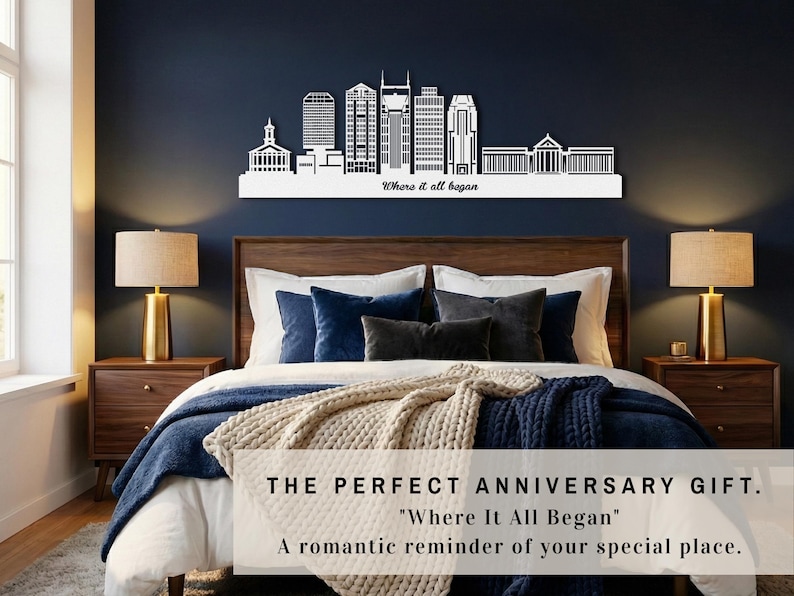 May include: A white skyline silhouette with the text "Where it all began" above a bed with a wooden headboard. The room has a navy blue wall, two bedside tables with lamps, and a cosy bedspread.