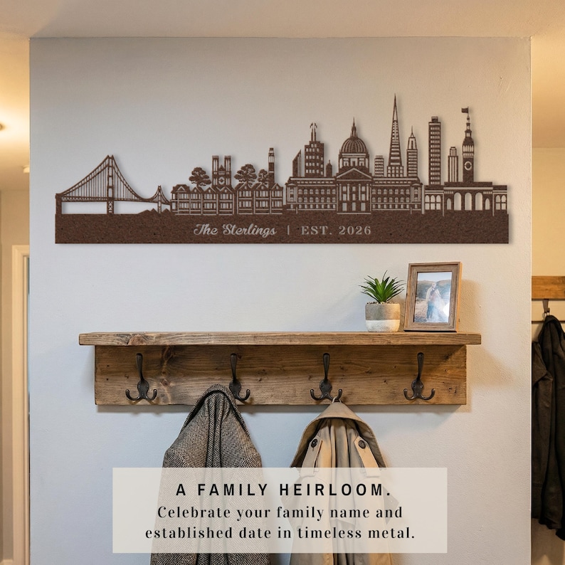 May include: A brown metal skyline wall art piece with the text "The Sterlings | EST. 2026". Below, a wooden shelf with hooks holds coats. A framed photo and a potted plant are also on the shelf. The text at the bottom reads "A FAMILY HEIRLOOM."