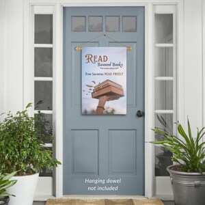 Może przedstawiać: Kolorowy baner na drzwi z tekstem "READ Banned Books Free Societies READ FREELY" i ilustracją dłoni trzymającej stos książek. Baner jest zawieszony na drewnianej listwie.