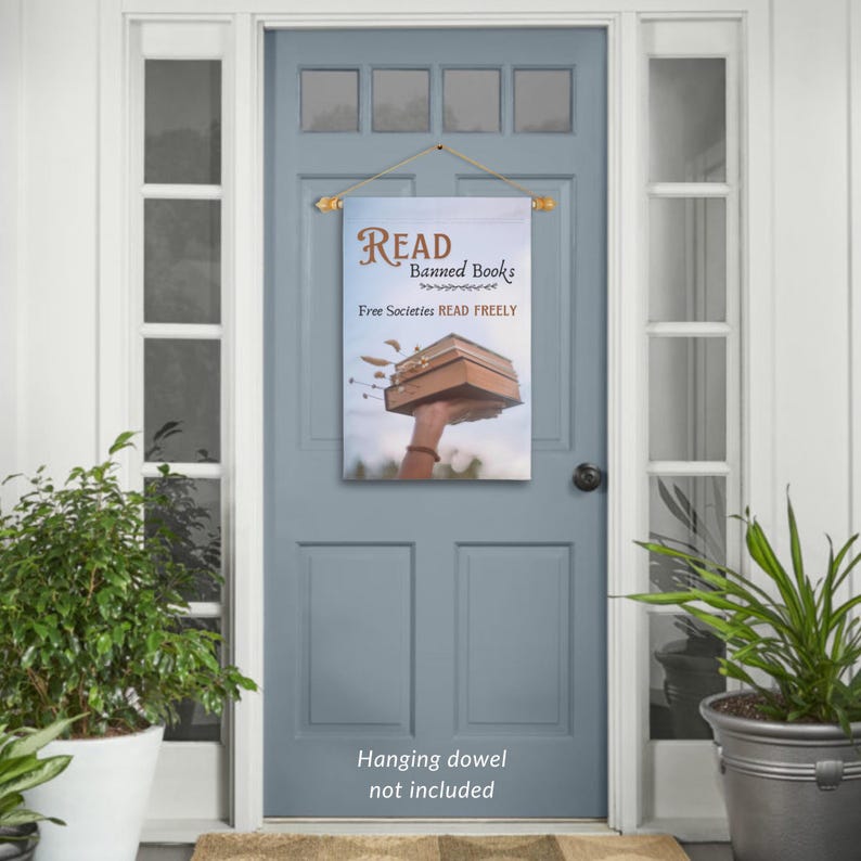 Może przedstawiać: Kolorowy baner na drzwi z tekstem "READ Banned Books Free Societies READ FREELY" i ilustracją dłoni trzymającej stos książek. Baner jest zawieszony na drewnianej listwie.