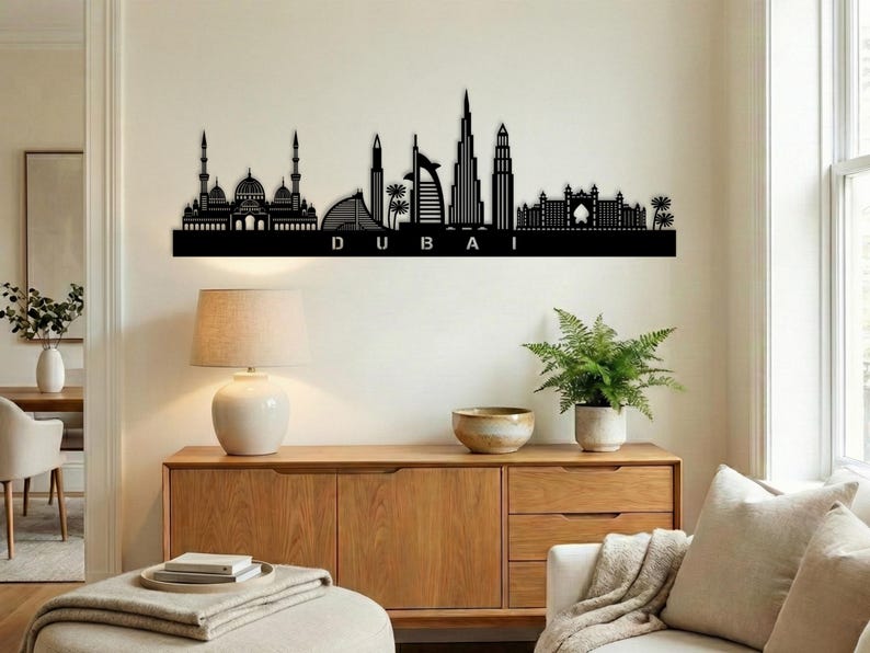 May include: Black metal wall art depicting the Dubai skyline, including iconic buildings and structures. The word "DUBAI" is displayed in capital letters below the skyline. The artwork is mounted on a white wall above a wooden cabinet.