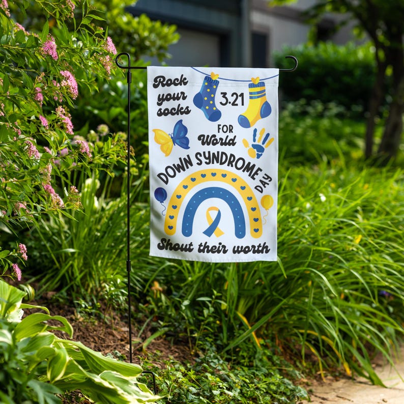 K&ouml;nnte beinhalten: Eine wei&szlig;e Gartenflagge mit einem blauen und gelben Regenbogen, einem gelben Band und dem Text "Rock your socks for World Down Syndrome Day. Shout their worth." Die Flagge zeigt au&szlig;erdem einen Schmetterling, einen Handabdruck und zwei Socken.