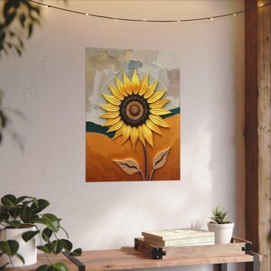 Puede incluir: Una impresi&oacute;n art&iacute;stica enmarcada que presenta un gran girasol texturizado. El girasol tiene p&eacute;talos de color amarillo brillante y un centro marr&oacute;n oscuro. El fondo incluye tonos de naranja, verde y gris, creando una escena c&aacute;lida. La obra de arte se exhibe en una pared.