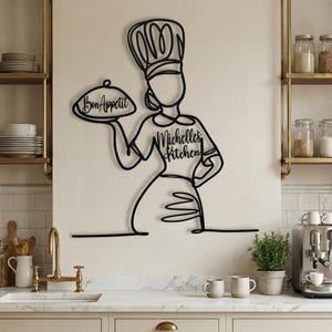 Black personalized metal kitchen sign reading "Michelle's Kitchen" mounted on a beige wall above a farmhouse sink and gold faucet.