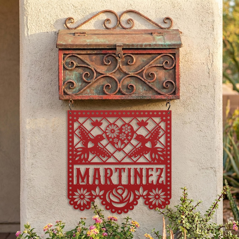 Op de afbeelding: Rood metalen bord met de naam "MARTINEZ" uitgesneden, met kolibries en bloemmotieven. Het bord hangt onder een verweerde, sierlijke brievenbus met een koperen afwerking. De brievenbus en het bord zijn op een stucwerkmuur gemonteerd.