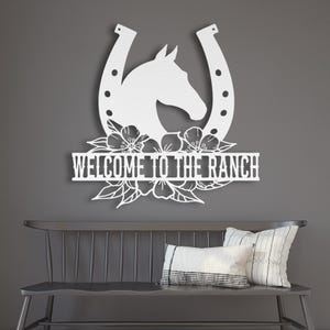 May include: White metal wall art featuring a horse head within a horseshoe, with the text "WELCOME TO THE RANCH" below. Floral accents surround the text. The artwork is displayed on a gray wall above a bench with pillows.