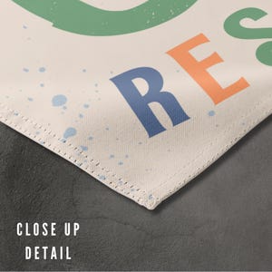 May include: Close-up detail of a white canvas with a stitched edge. The canvas has a blue, orange, and green design with the word "REST" printed in large letters.