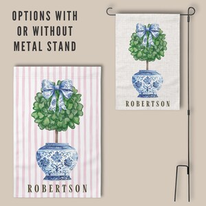May include: A personalized garden flag with a blue and white porcelain vase with a green topiary tree and a blue bow on top. The flag has pink and white stripes and the text "ROBERTSON" at the bottom.