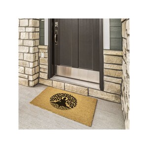 May include: A brown coir doormat with a black silhouette of a person sitting in a tree pose with roots extending from the tree. The doormat is in front of a brown door with a silver door kick plate.
