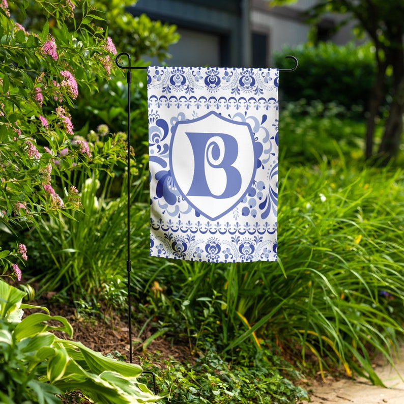 May include: A blue and white garden flag with a white shield featuring the letter "B" in blue. The flag has a blue and white floral pattern.