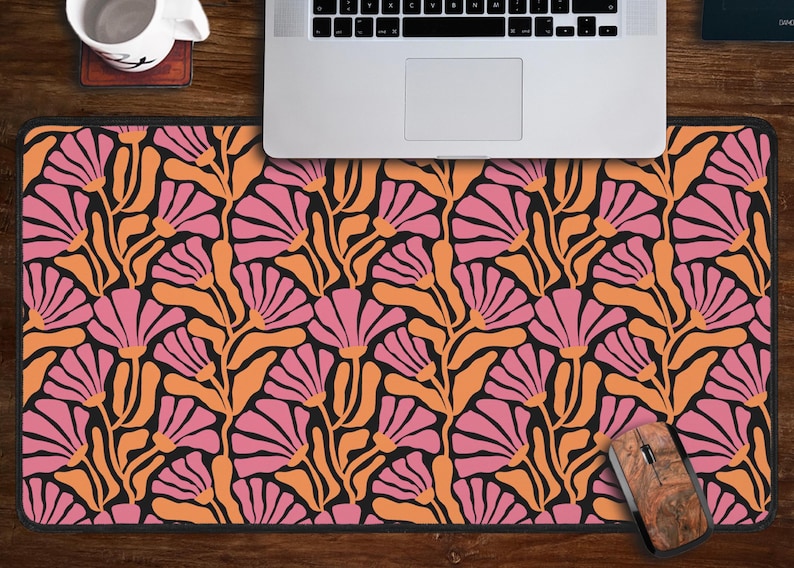May include: A pink and orange floral patterned mouse mat with a black background. The mouse mat is on a wooden desk with a laptop and a mouse.