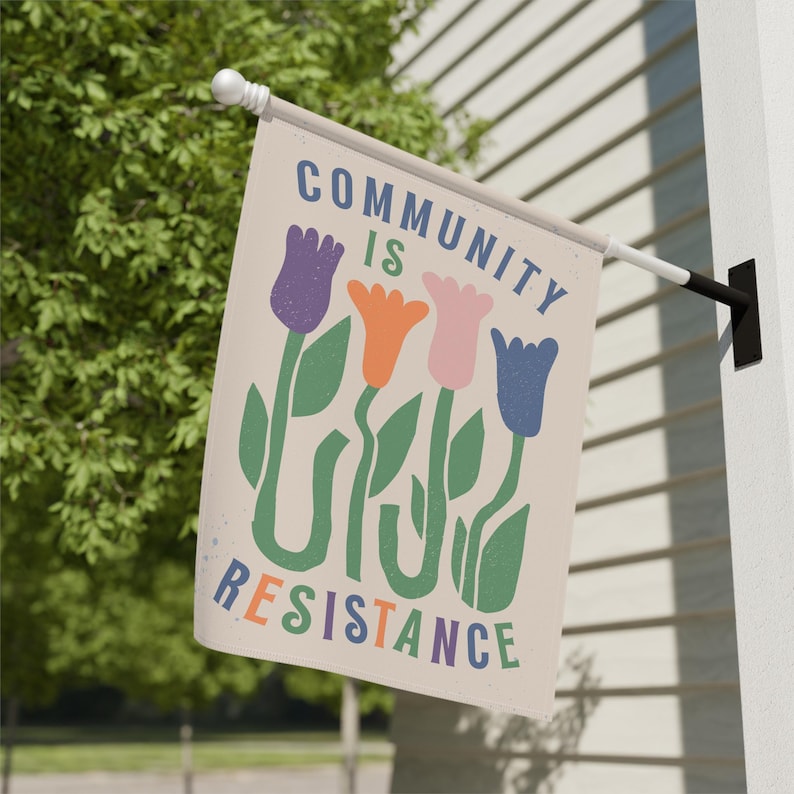 May include: A beige flag with the text "Community is Resistance" in colourful letters. The flag features a stylised design of four tulips in purple, orange, pink, and blue.
