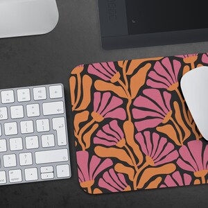 May include: A black mouse mat with a repeating pattern of pink and orange stylised flowers. The mouse mat is on a grey surface with a keyboard and a tablet.