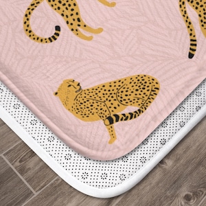 May include: A pink bath mat with a white non-slip backing features a repeating pattern of yellow cheetahs. The cheetahs are in various poses, some are lying down, some are standing, and some are walking.