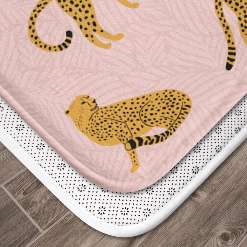 May include: A pink bath mat with a white non-slip backing features a repeating pattern of yellow cheetahs. The cheetahs are in various poses, some are lying down, some are standing, and some are walking.