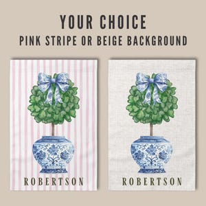 May include: Two garden flags with a blue and white porcelain pot with a green topiary tree and a blue bow. The flag on the left has a pink and white striped background and the flag on the right has a beige background. Both flags have the name "ROBERTSON" printed at the bottom.