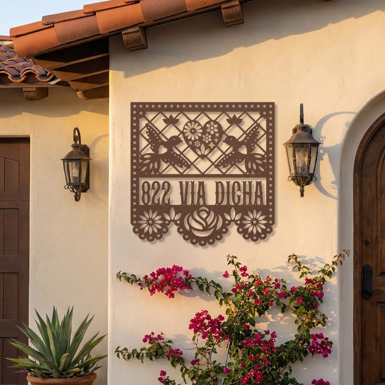 Op de afbeelding: Een bruin metalen adresbord met de tekst "822 VIA DICHA" is bevestigd aan een stucwerkmuur. Het bord heeft een decoratief uitgesneden ontwerp met vogels, bloemen en geometrische patronen. Een bloeiende klimplant omgeeft het bord.
