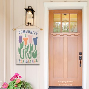 May include: A beige canvas flag with the text "Community is Resistance" in black lettering. The flag features a stylised design of four colourful tulips with green stems and leaves.